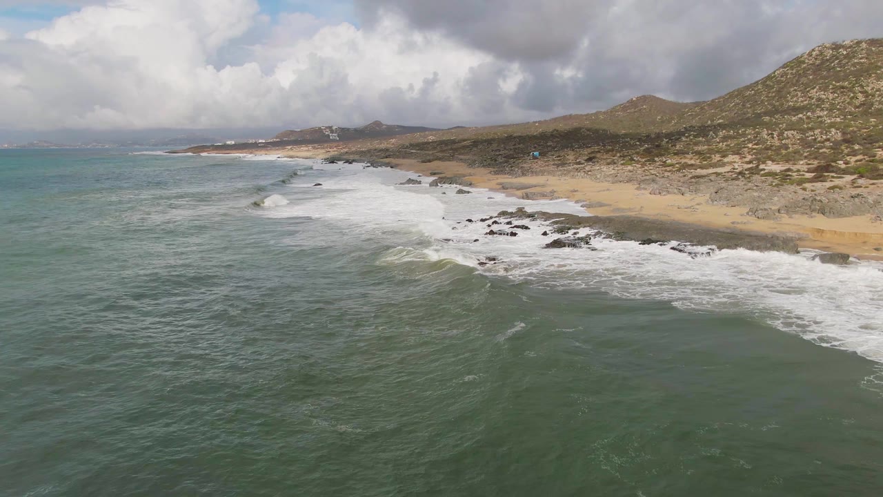 At 0:02, the wave line has advanced across the midground water toward the rocky Punta Gorda beach, with tan sand, brown-green hills, and no visible people in frame.