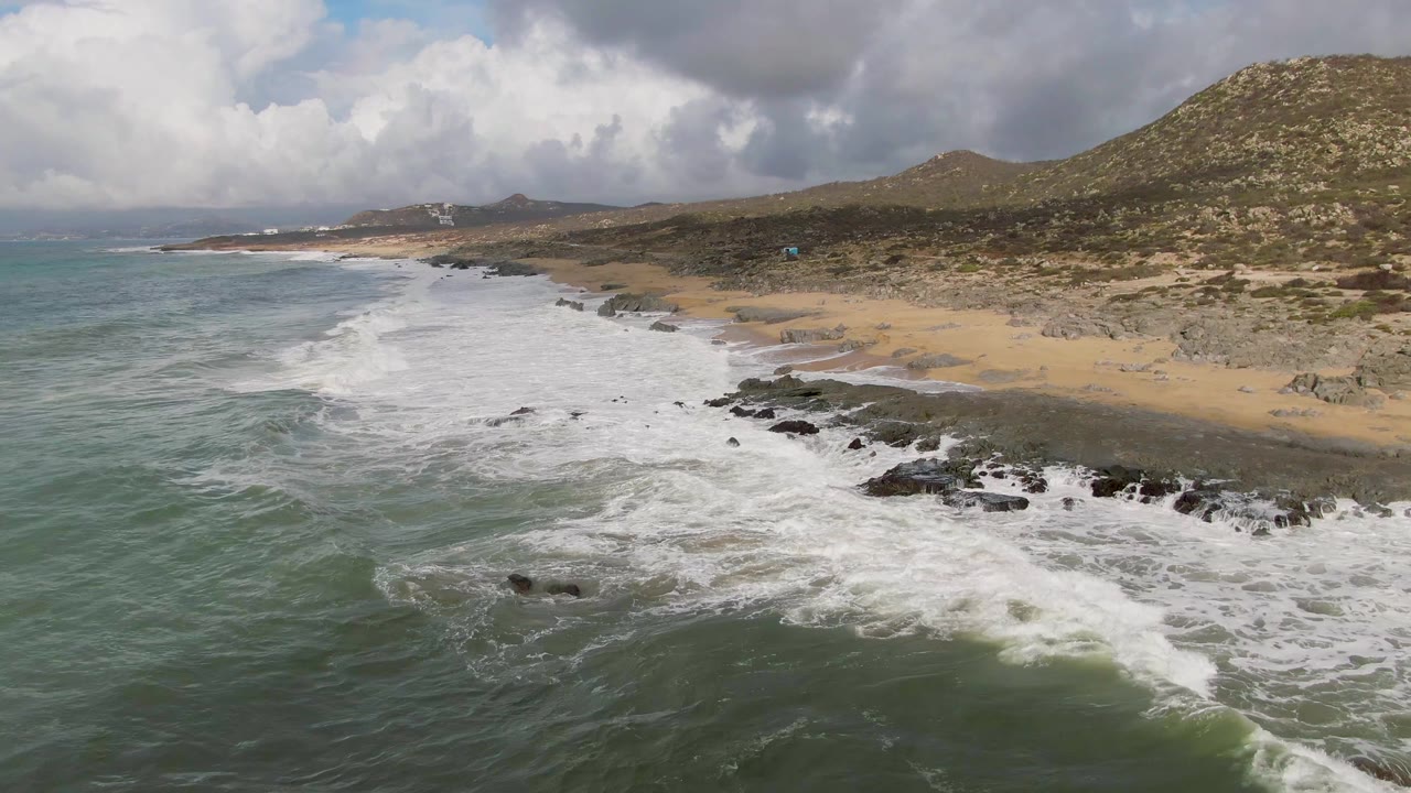 Aerial frame from Punta Gorda, Baja California Sur showing green-blue water pushing white surf over dark rocks toward a tan beach, with scrub hills behind and no visible people.