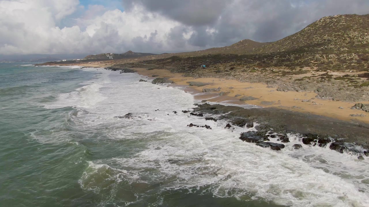 Aerial view in Punta Gorda, Baja California Sur with a broad band of whitewater crossing the rocky shore, tan sand to the right, hills beyond, and no visible people.