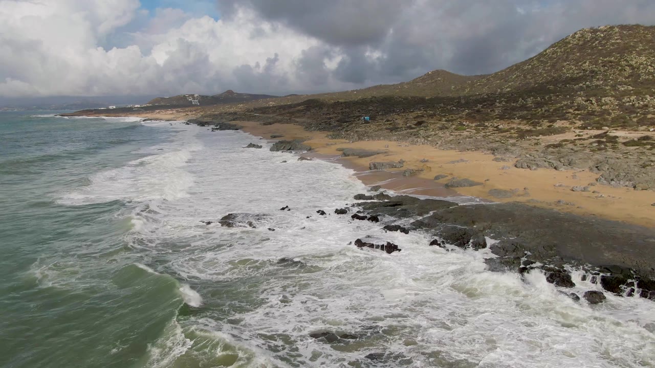 Punta Gorda, Baja California Sur shoreline seen from above at an angle, with surf curling in from the left, foam around black rocks, open sand to the right, and no visible people.
