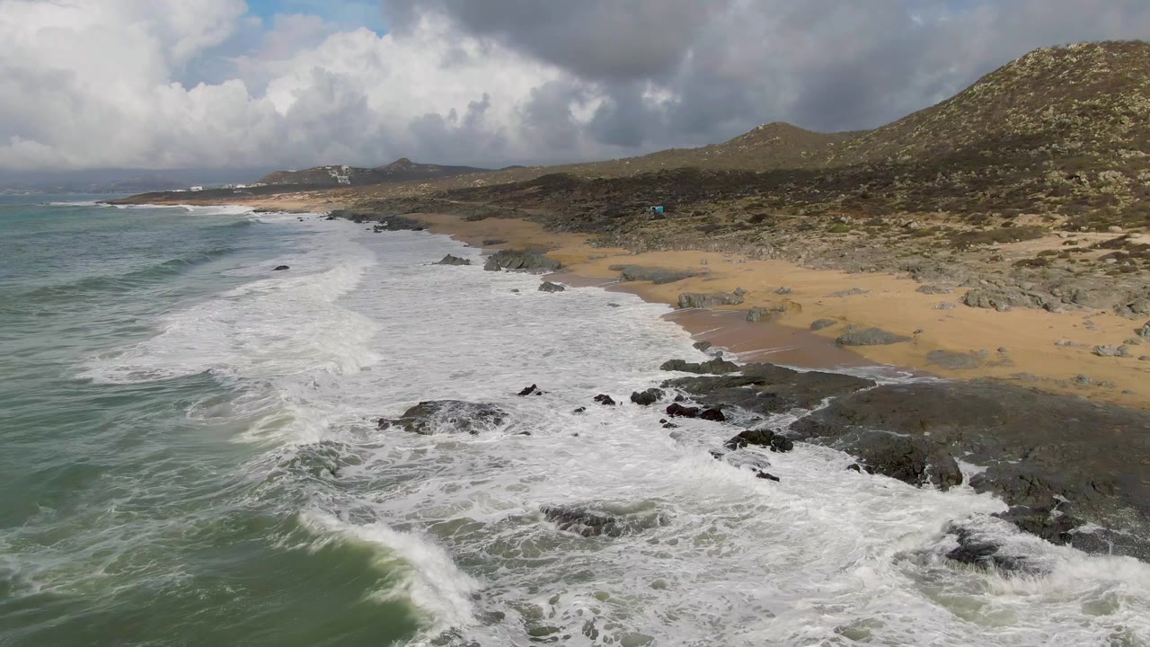 Aerial coastline with foamy surf, scattered rocks, beach, and hills