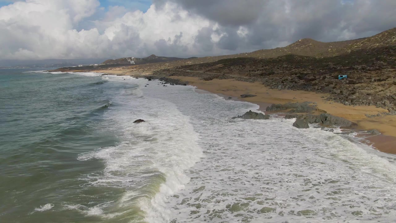Aerial view of waves rolling toward a sandy beach with rocks and hills