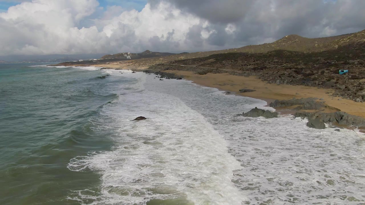 At 0:01, the Punta Gorda shoreline fills the frame with a broad band of white foam, green-blue water at left, sand at right, rocky outcrops, hills, and no visible people.