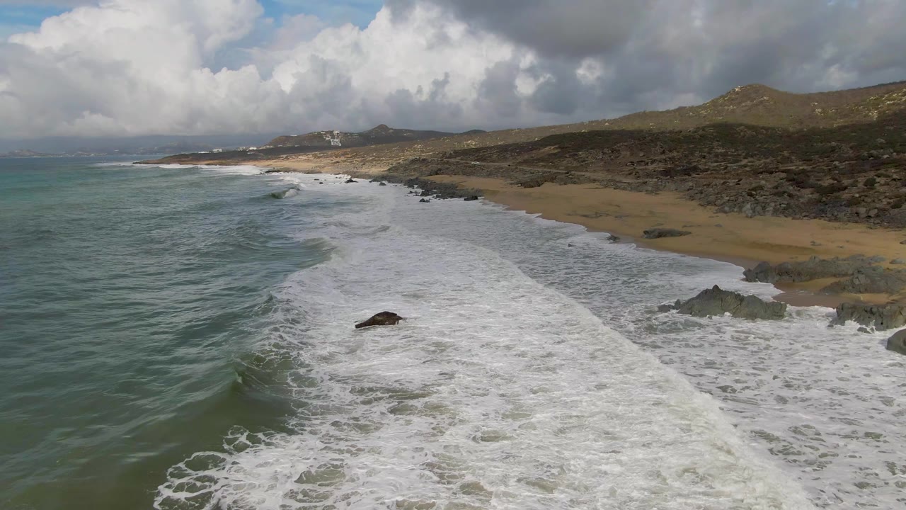 At 0:02, breaking waves wrap around darker rocks near the sandy beach at Punta Gorda, with textured water, arid hills, cloud shadow, and no visible people.