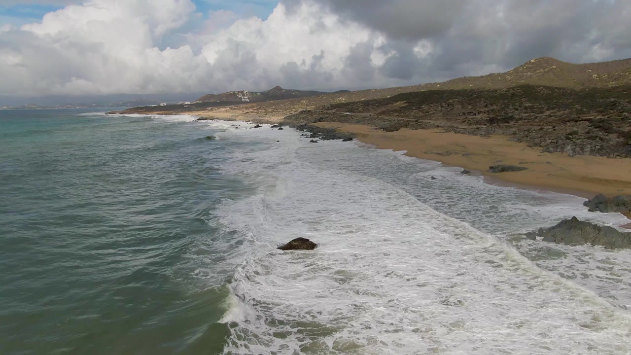 At 0:03, the aerial view looks along the Punta Gorda coast with ocean on the left, a long foam band near center, sandy beach and rocks on the right, hills beyond, and no visible people.