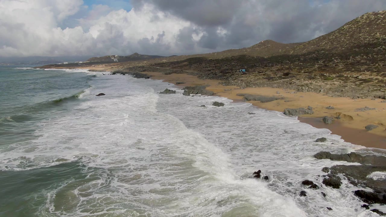 Aerial frame from Punta Gorda, Baja California Sur, showing foamy surf entering from the left toward tan sand, scattered dark rocks, low hills, cloudy sky, and no visible people.