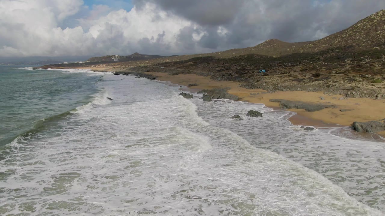 Aerial frame from Punta Gorda, Baja California Sur, showing surf pushed farther across the frame, with exposed sand and rocks on the right, darker hillside terrain behind, cloud shadows, and no visible people.