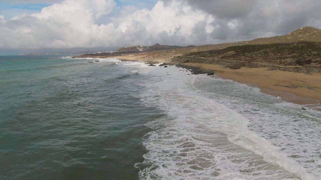Aerial view of Punta Gorda, Baja California Sur, with white surf spreading across teal water toward a sandy beach, dark rocks along the shore, hills in the distance, and no visible people.