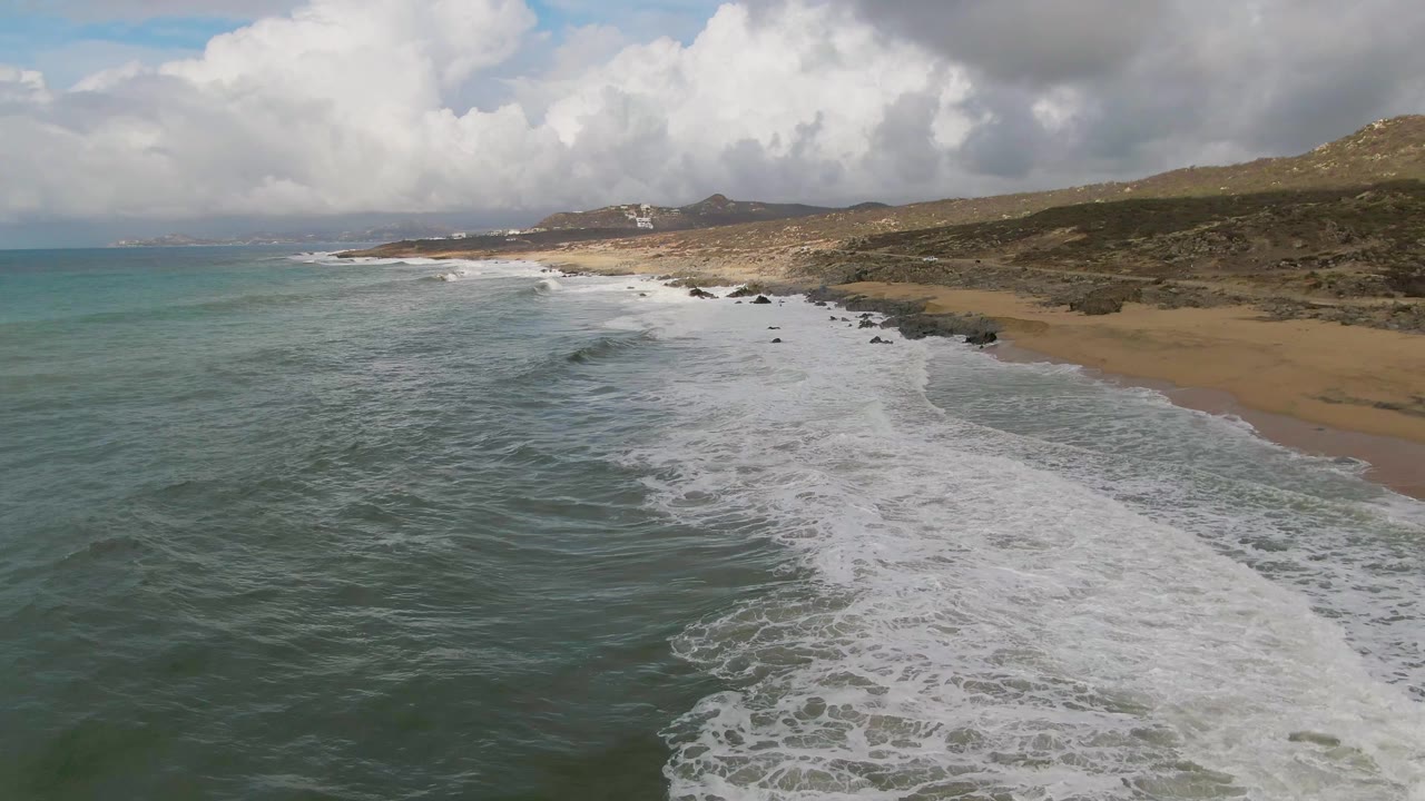 Aerial frame over Punta Gorda showing a broad band of white foam advancing diagonally toward the sand, with turquoise water on the left, rocky shoreline on the right, distant hills, and no visible people.