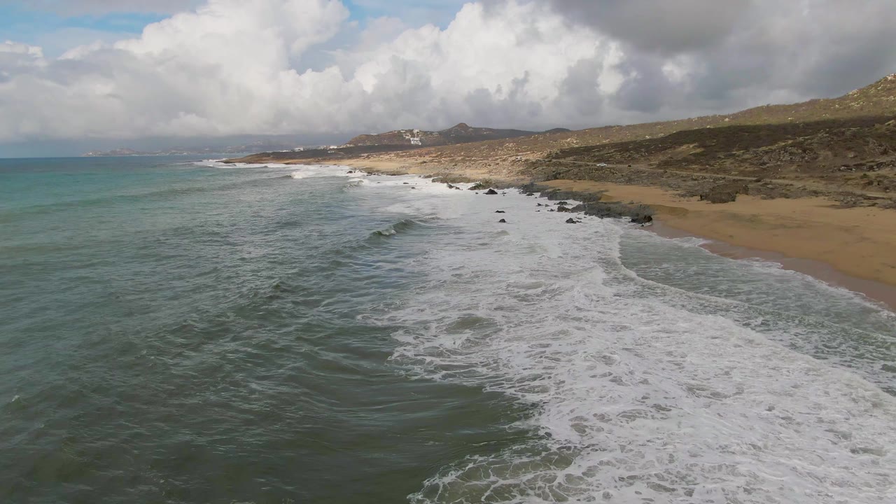Aerial view of Punta Gorda coastline where a smaller wave forms offshore before merging into the wider foam field, with sand, scattered rocks, hills, cloudy sky, and no visible people.