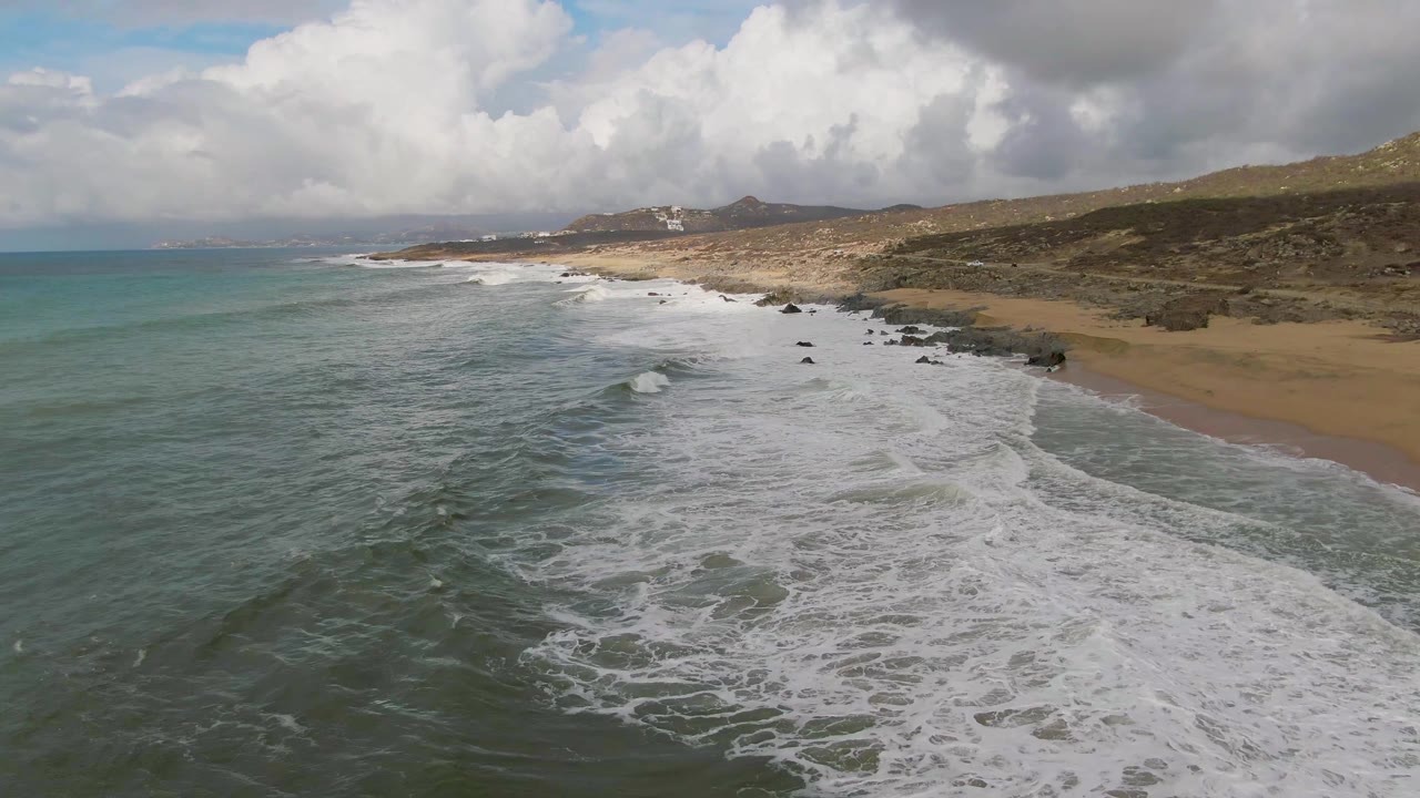 Aerial frame at Punta Gorda with the surf spread wider across the foreground, open ocean filling the left side, sandy beach and rock clusters running along the right, distant hills, and no visible people.
