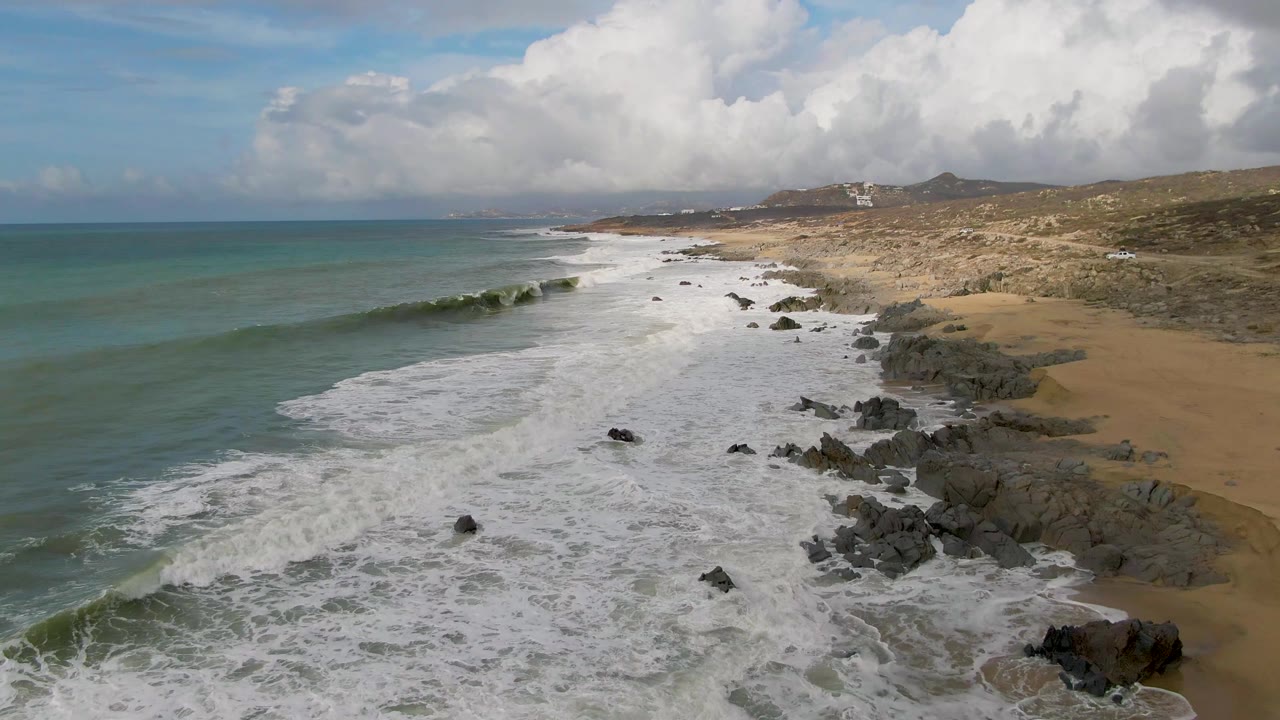 Aerial view of Punta Gorda surf spreading toward dark shoreline rocks and tan sand, with green-blue ocean to the left, scrubland and hills beyond the beach, and no visible people.