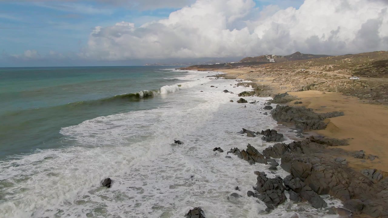 Aerial Punta Gorda coastline view with a green wave starting to curl near offshore rocks, white foam spread across the beach edge, dry coastal terrain inland, and no visible people.