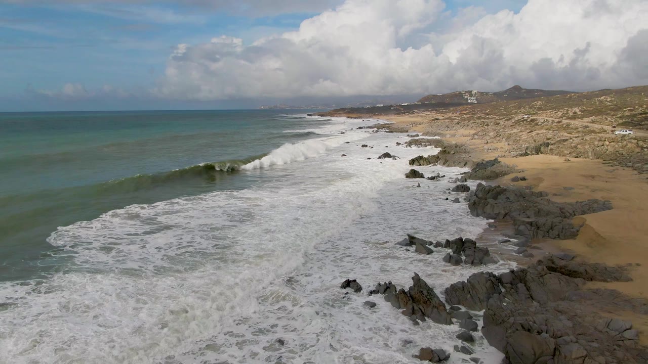 Aerial frame from Punta Gorda showing the wave breaking into bright white spray over rocks, with foamy water filling the shore, sandy beach to the right, and no visible people.
