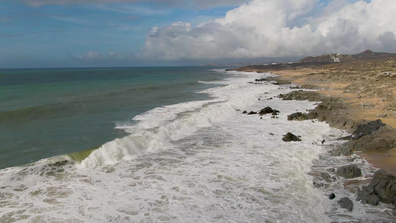 At Punta Gorda in Baja California Sur, white surf breaks across rocks and sand with green-blue ocean to the left, coastal hills in the distance, and no visible people.
