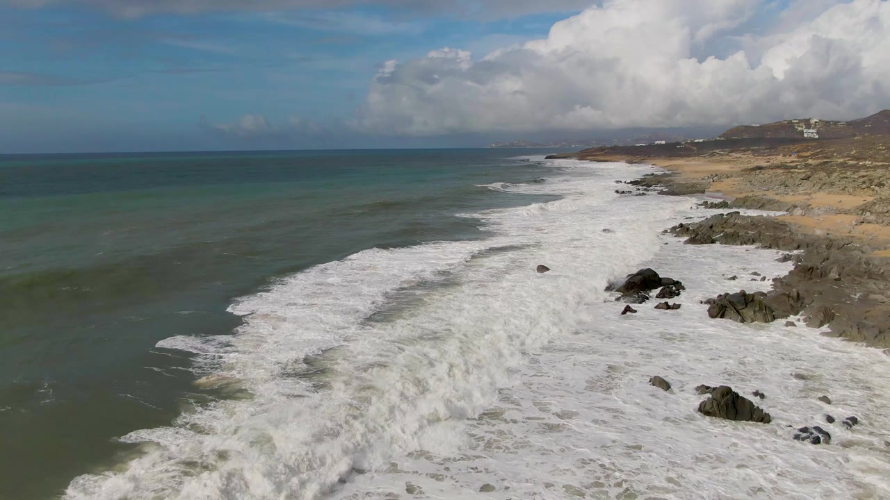 At Punta Gorda in Baja California Sur, foamy water spreads farther along the shoreline, with exposed rocks, pale beach, distant hillside structures, and no visible people.