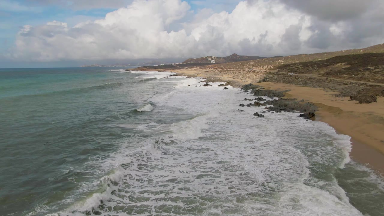 At 0:01, the Punta Gorda shoreline remains angled across the frame as fresh white foam spreads farther over the nearshore water, with beach, rocks, low hills, and no visible people.