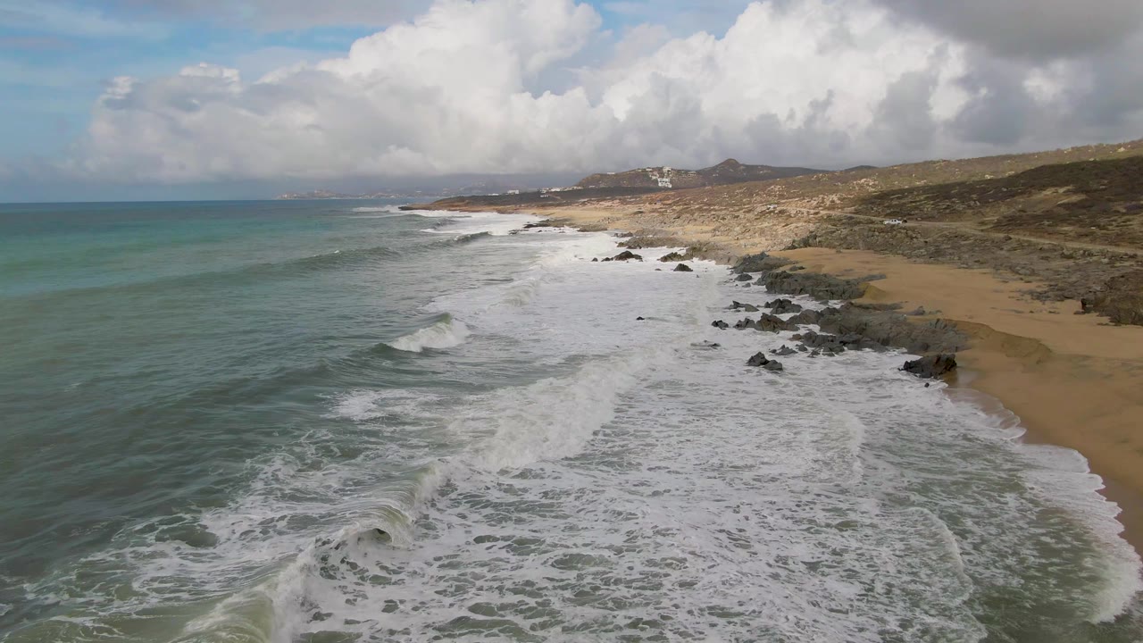 At 0:02, white water covers more of the rocky nearshore at Punta Gorda while deeper blue ocean sits to the left, the sand curves along the right side, and no visible people appear.