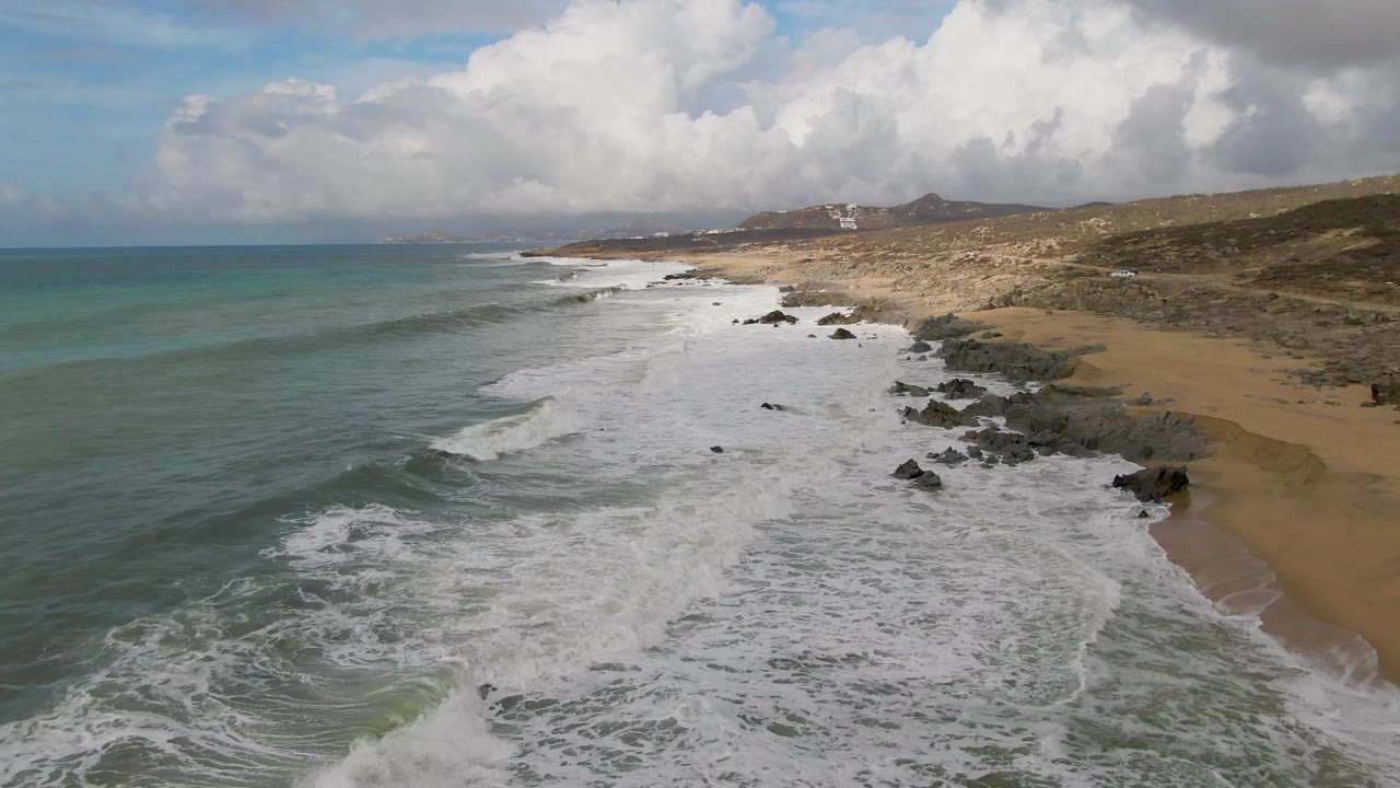 At 0:03, a cleaner wave line forms in the left foreground while broken foam fills the shoreline at Punta Gorda, with tan sand, dark rocks, hills, clouds, and no visible people.