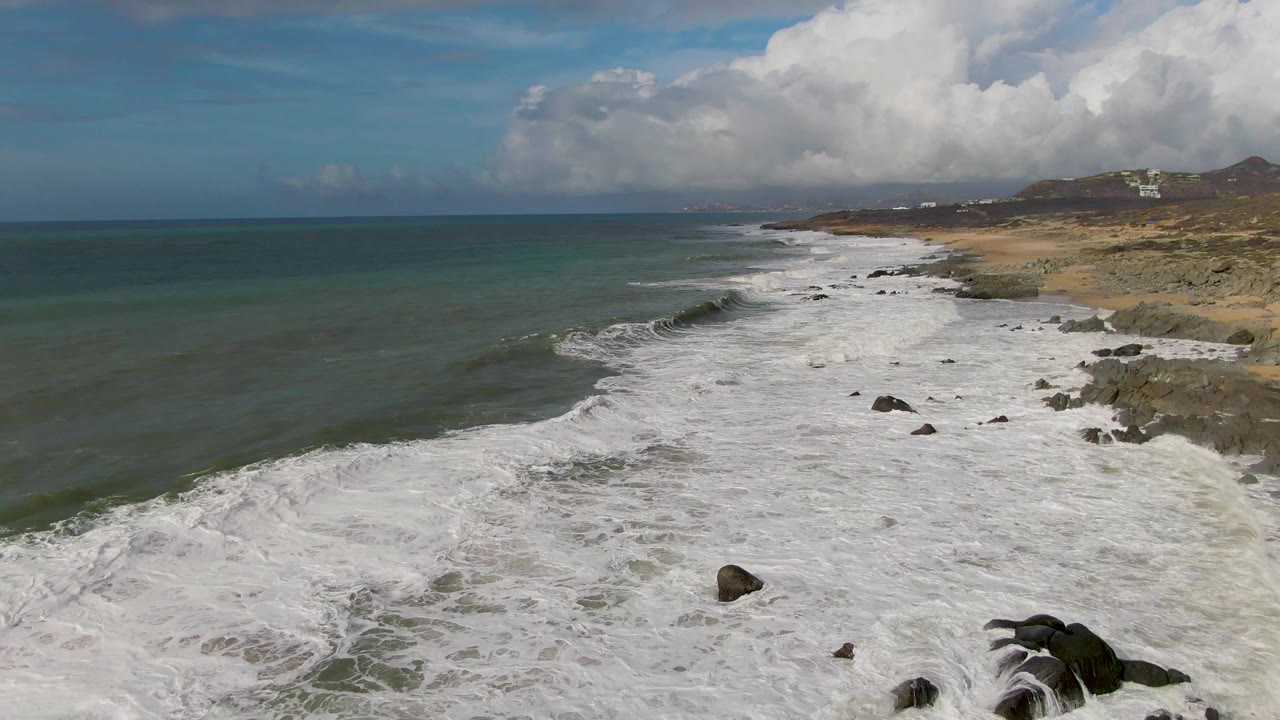 Aerial view of Punta Gorda East Cape surf spreading across a rocky sandy shoreline, with green-blue ocean to the left, distant hills under clouds, and no visible people.