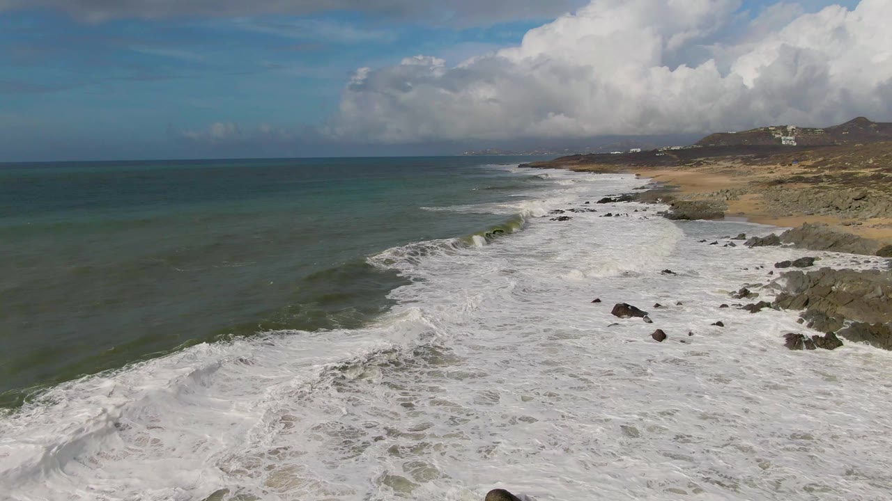 Foamy waves roll toward rocks and sand below a cloudy coastal sky.