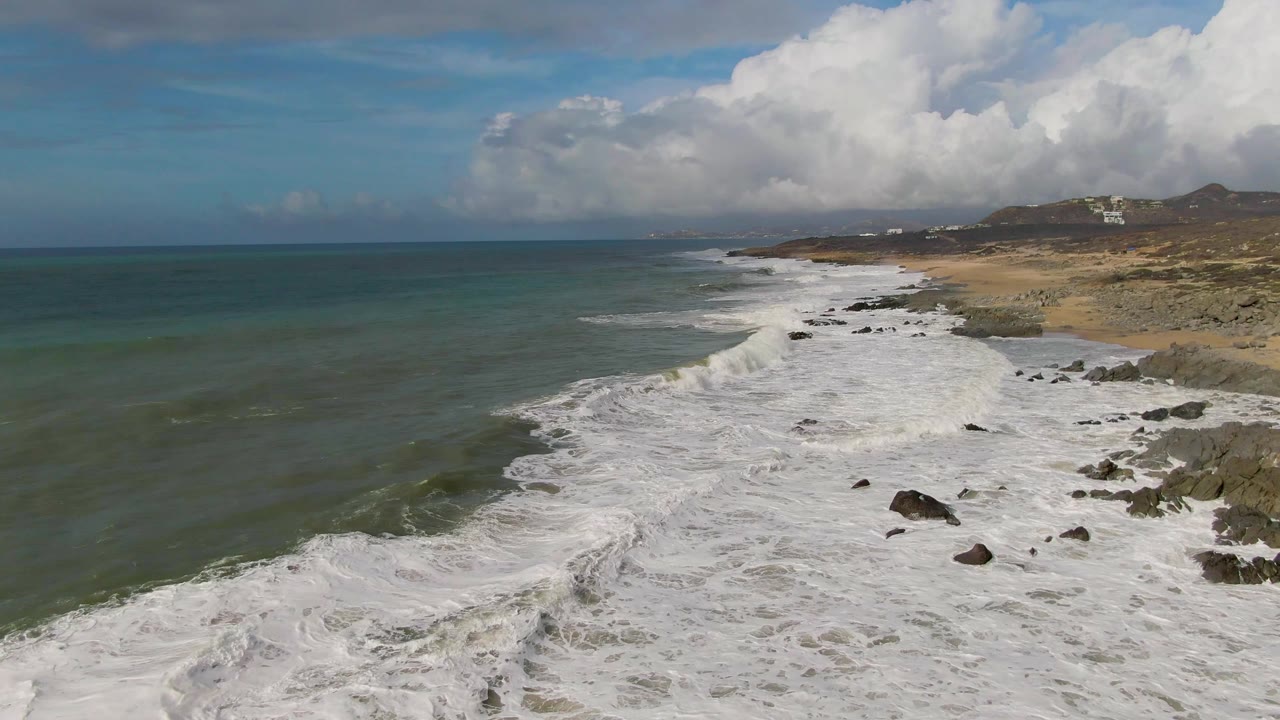 Whitewater curls around dark rocks on the Punta Gorda East Cape coast, with the sandy shore and low hills continuing into the distance and no visible people.