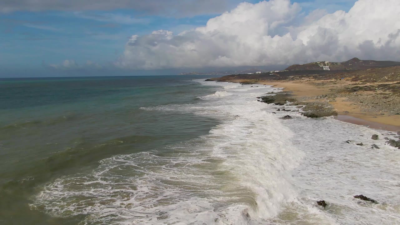 Aerial view of Punta Gorda East Cape surf rolling toward a sandy and rocky shore, with green-blue water on the left, low hills in the distance, and no visible people.