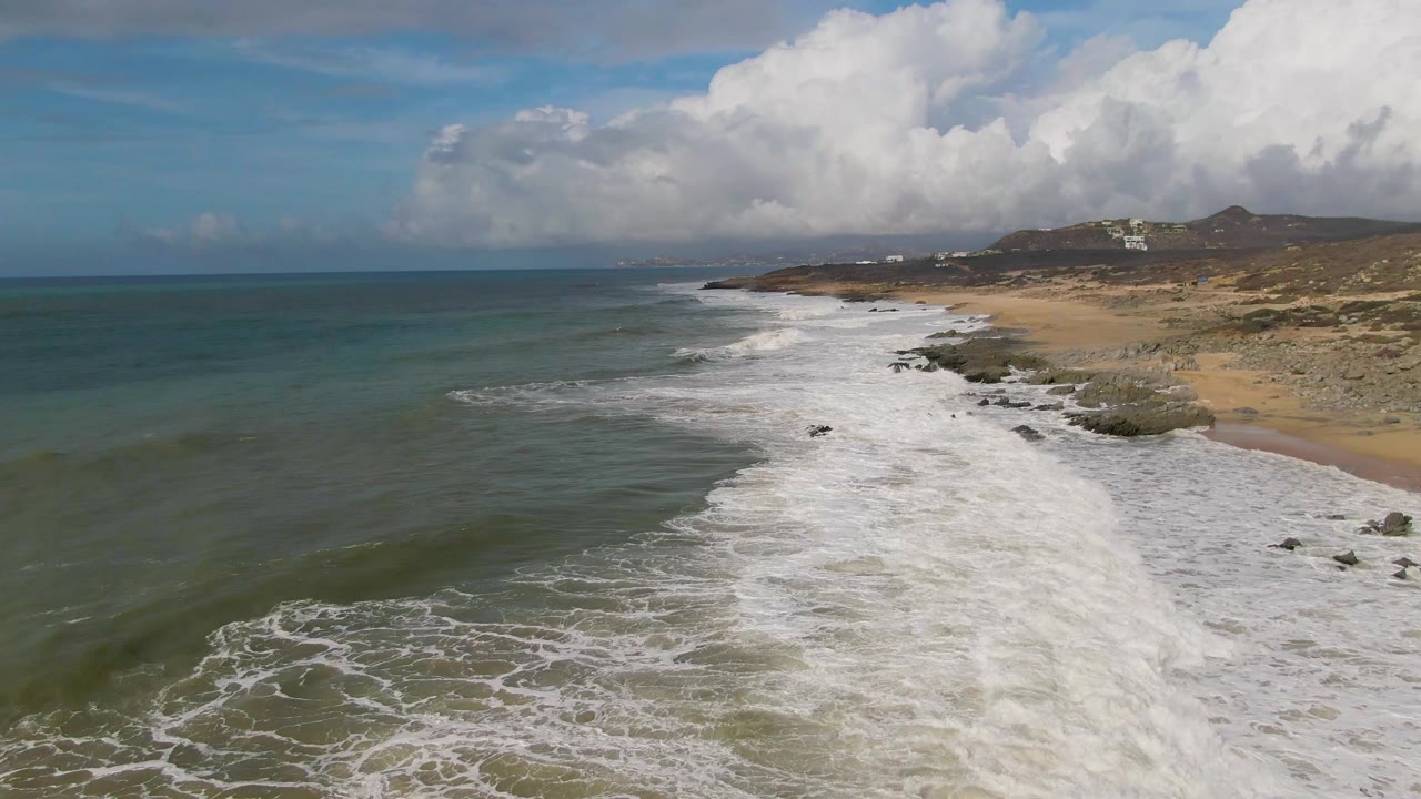 White surf spreads across a sandy beach with rocks and hills beyond.