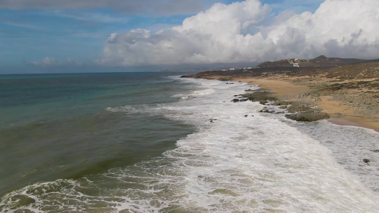 Aerial view at Punta Gorda East Cape with surf wrapping around scattered rocks, a tan beach and dry hillside to the right, open sea to the left, and no visible people.