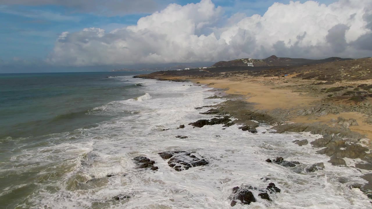 A broad band of whitewater rolls into the rocky shore at Punta Gorda, with the sandy beach and low hills continuing into the background and no visible people.