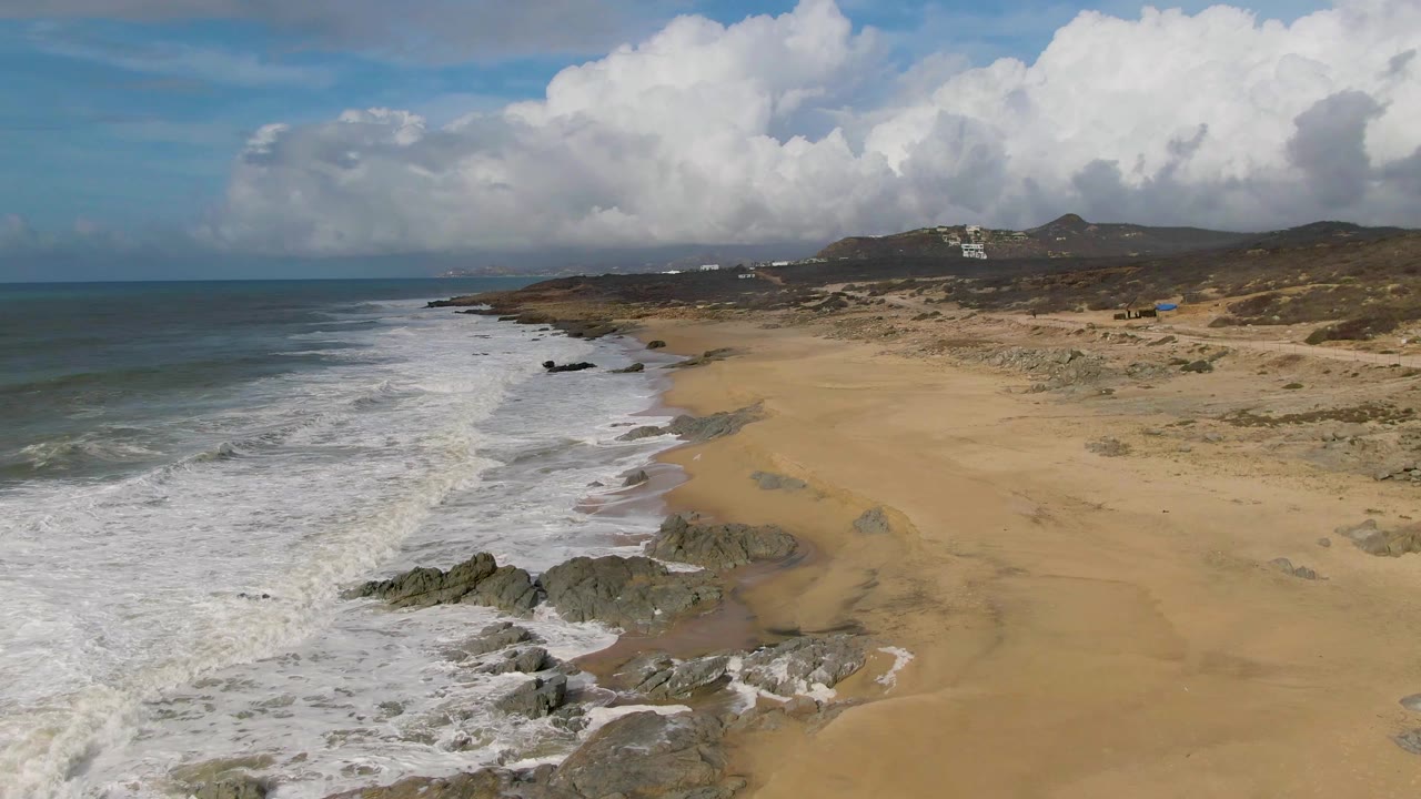 Aerial frame of Punta Gorda East Cape surf rolling onto a broad sandy beach with dark rocks in the foreground, dunes and hills beyond, and no visible people.