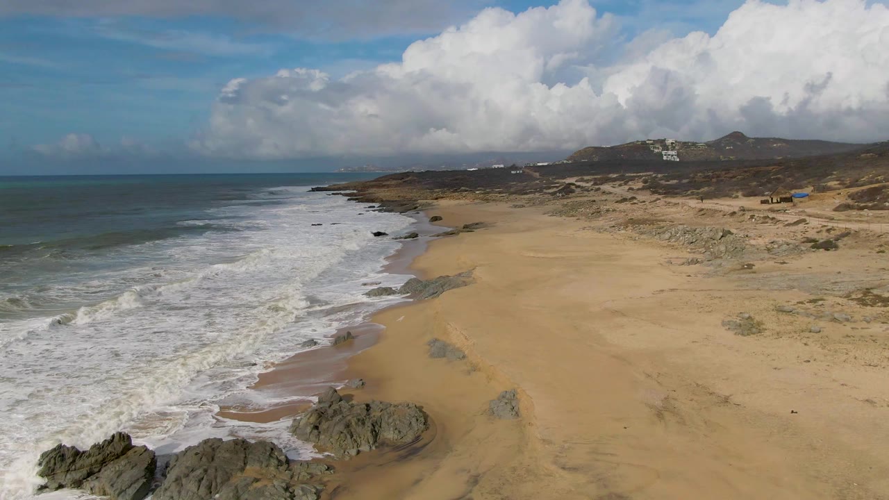 Aerial frame from Punta Gorda East Cape showing waves running diagonally along the beach, rocky outcrops near the lower edge, dune terrain inland, and no visible people.