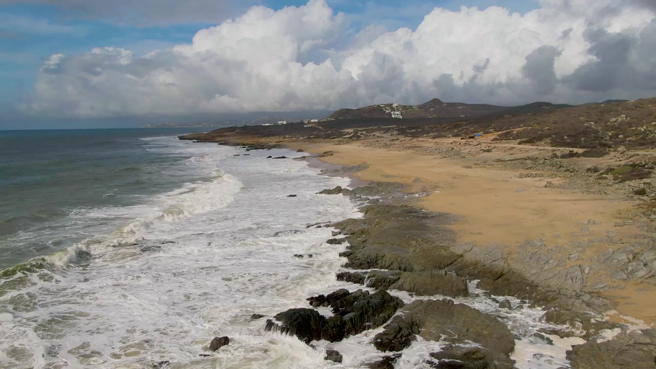 Aerial view of Punta Gorda, Baja California Sur, where white surf breaks over dark rocks beside tan sand, with low hills and no visible people.