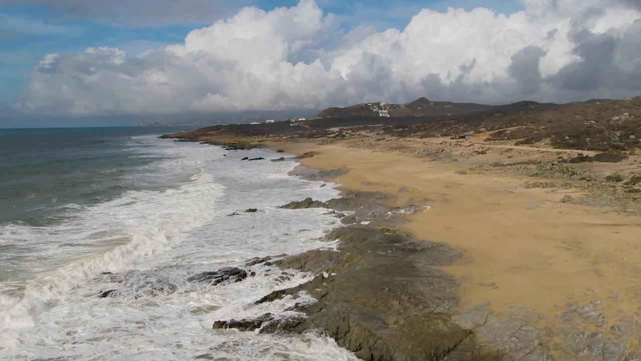 At Punta Gorda, surf lines angle into the rocky shore while the sandy beach opens to the right beneath large white and gray clouds, with no visible people.