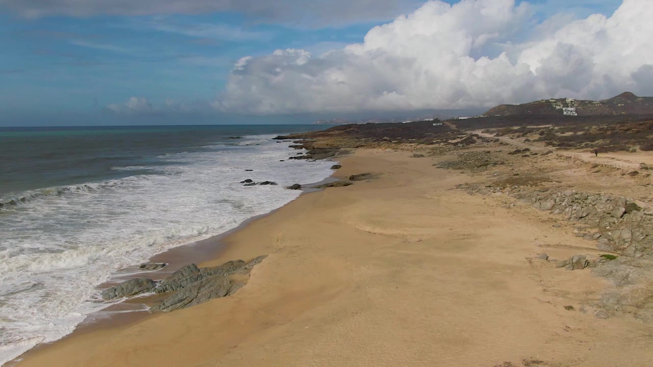 Aerial view at Punta Gorda East Cape, Baja California Sur, with white surf entering from the left, a broad sandy beach below, rocky outcrops near the waterline, clouds on the horizon, and no visible people.