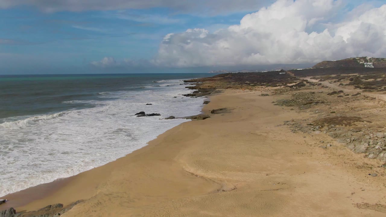At 0:02, the beach at Punta Gorda East Cape fills the foreground and center while waves curve in from the left, small rock formations break the surf, dunes and scrub run along the right side, and no visible people appear.