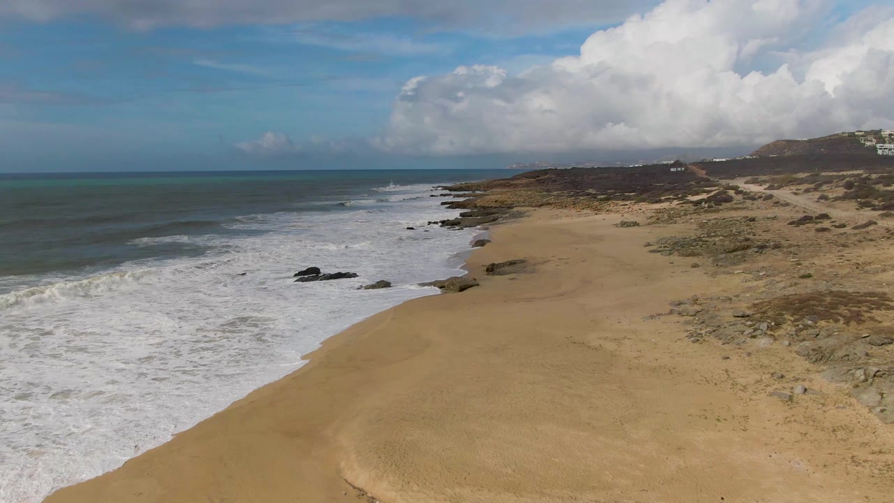 At 0:03, foamy water spreads across the left side of the frame, the sandy beach opens through the middle, rocky dune ground lines the right, distant clouds sit above the sea, and no visible people are present.