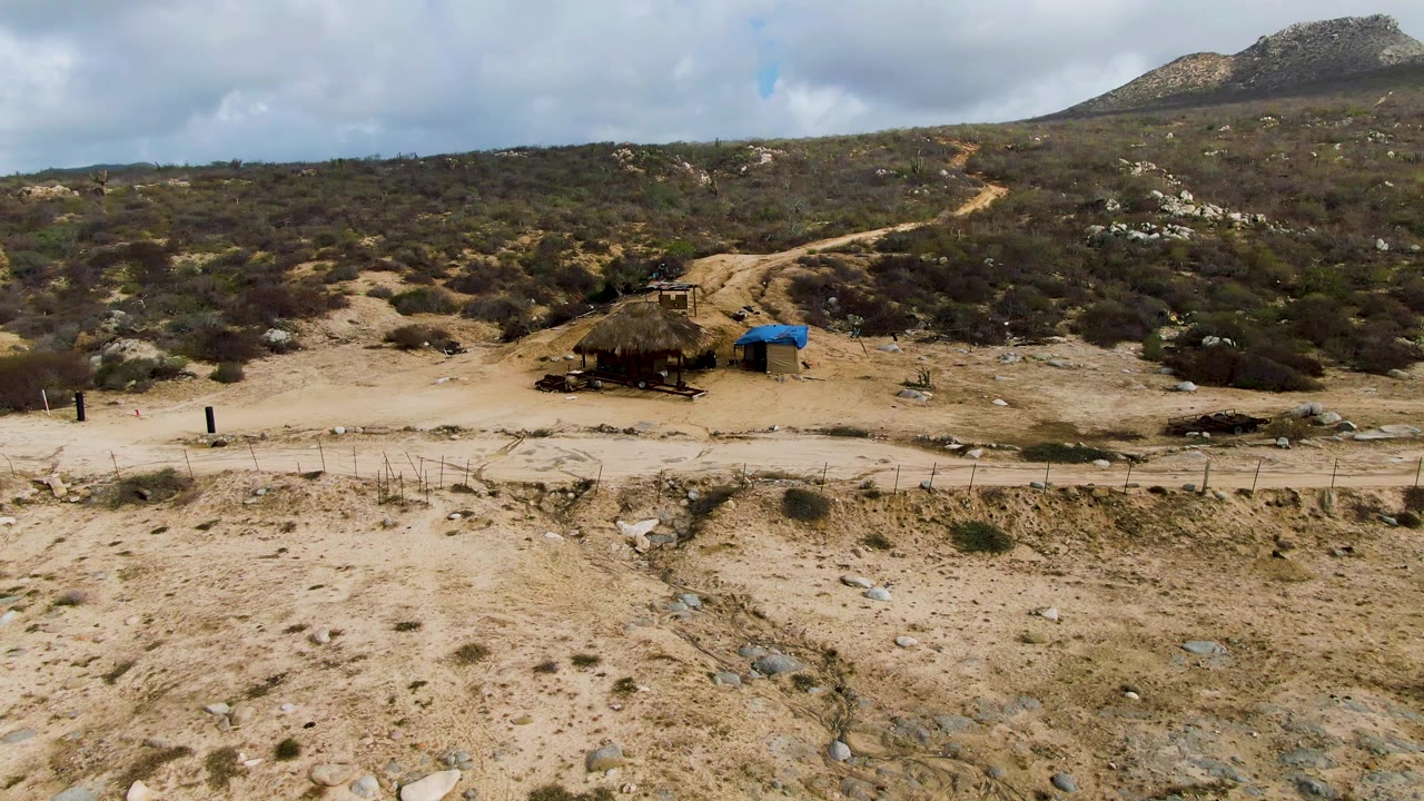Opening frame of a dirt road through Punta Gorda East Cape scrubland in Baja California Sur, with a small shade shelter, blue tarp, rocky hillside, cloudy sky, and no visible people.