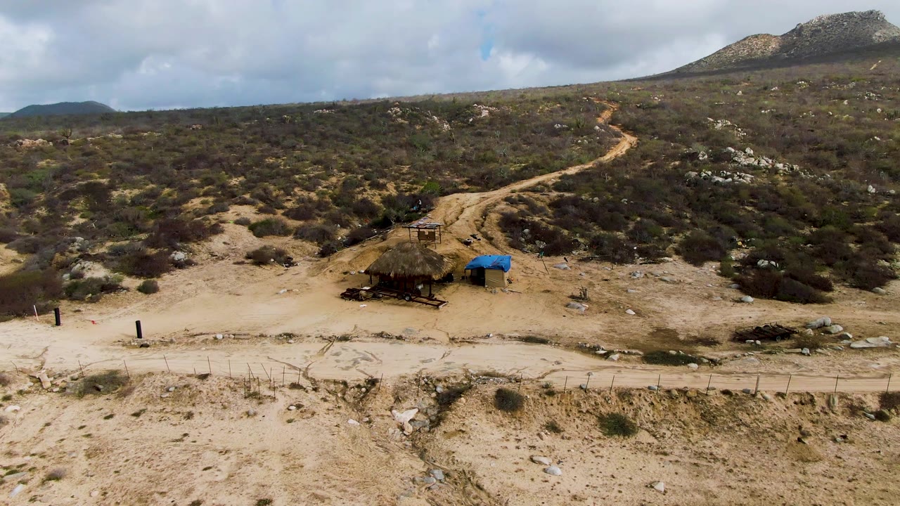 The road remains centered across the arid Punta Gorda hillside, with fence posts in the lower ground, the shelter and blue cover below the track, and no visible people.