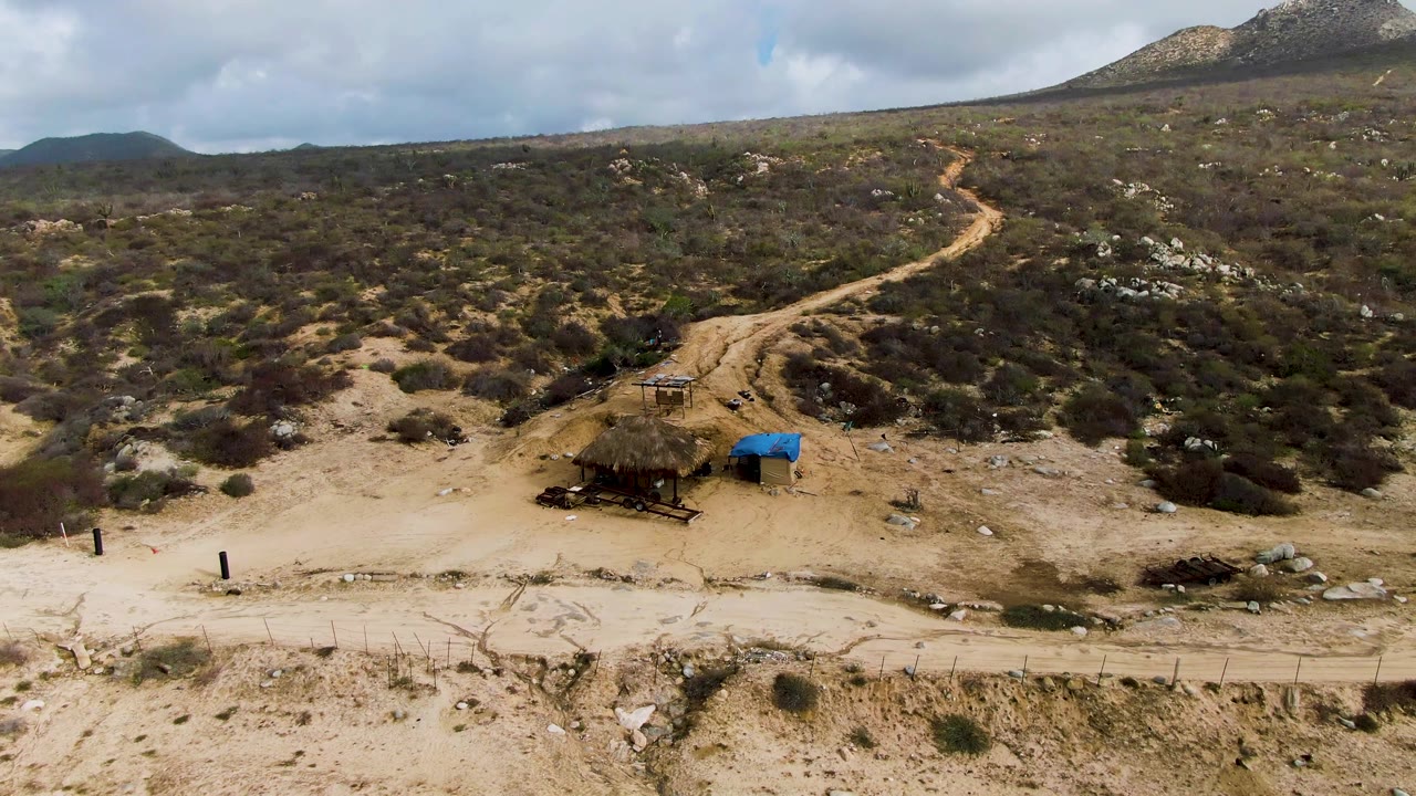 Dirt road and shelter viewed across dry terrain with scrub-covered hills behind.