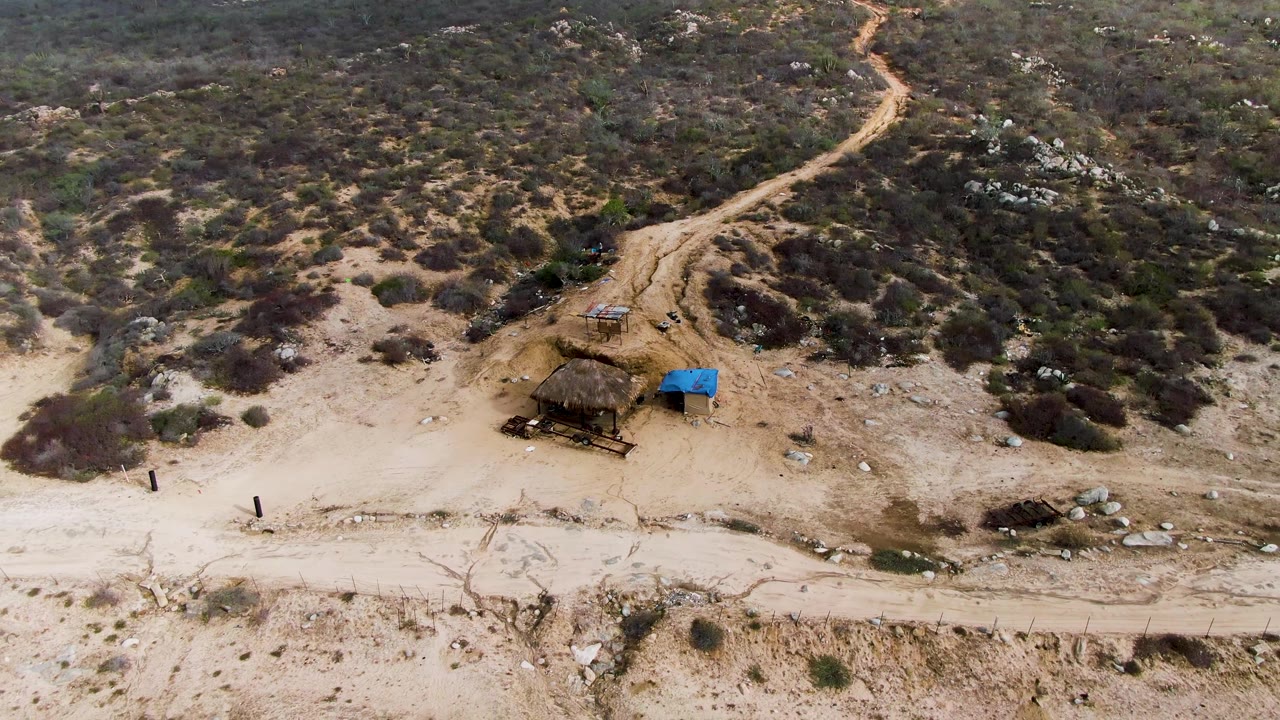 Aerial view in Punta Gordo, Baja California Sur, Mexico, showing a pale dirt road below scrub-covered terrain, a small rough shelter, a blue tarp, and tiny people near the road.