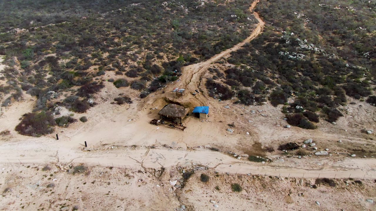 Aerial frame of dry bushland with a dirt road and blue-tarp structure.