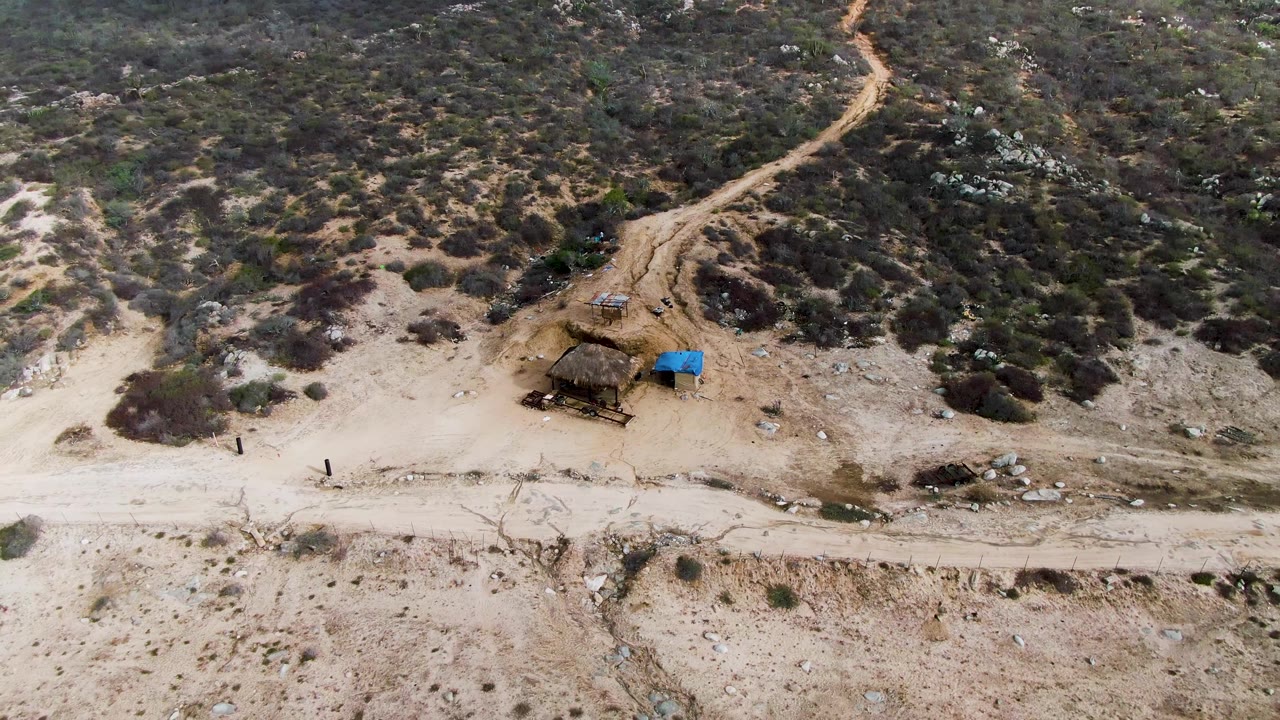 The frame shows Punta Gordo arid terrain from above, with a winding track rising through darker scrub, a pale road at the bottom, a small shelter, a blue tarp, and distant people near the roadside.