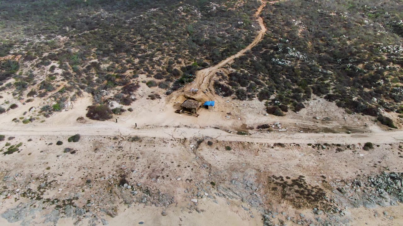 Aerial frame over Punta Gordo, Baja California Sur, showing pale sand, a parallel dirt road, a small rustic structure beside a blue tarp, scrubby hillside, and no visible people.