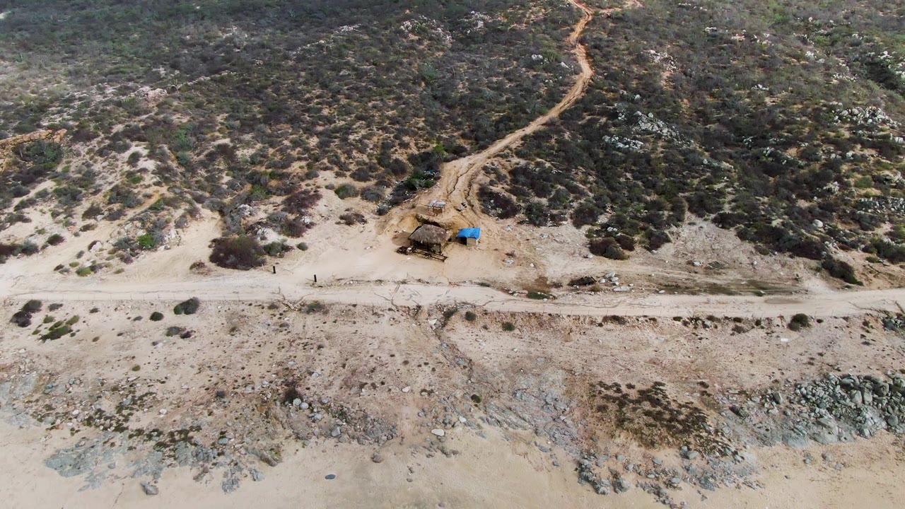 Aerial view at Punta Gordo with wet sand along the lower edge, pale dry beach, rocky outcrops near the road, scrub-covered slope, a branching track, and no visible people.