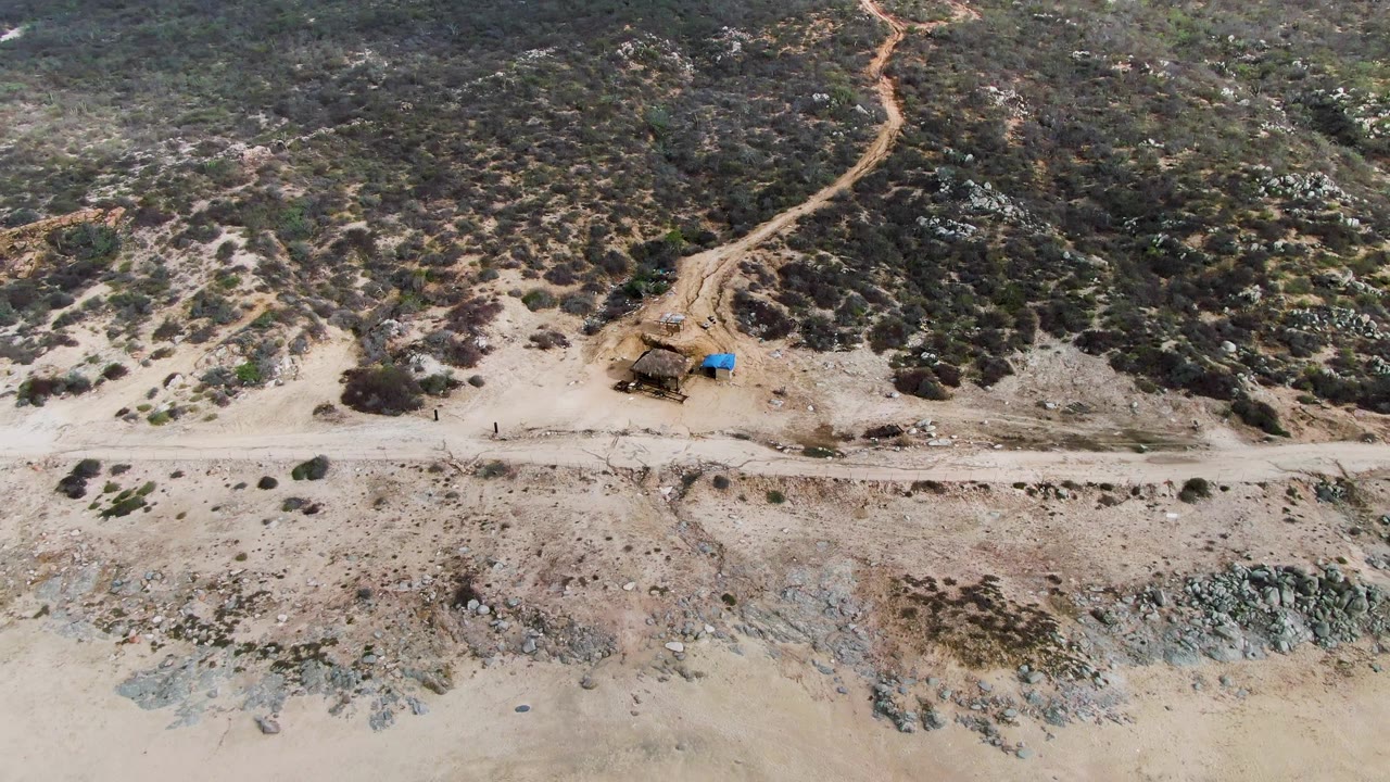 Drone frame over Punta Gordo's East Cape coast showing a broad exposed sand band, a road running parallel to the shore, uneven rocky ground, desert scrub, and no visible people.