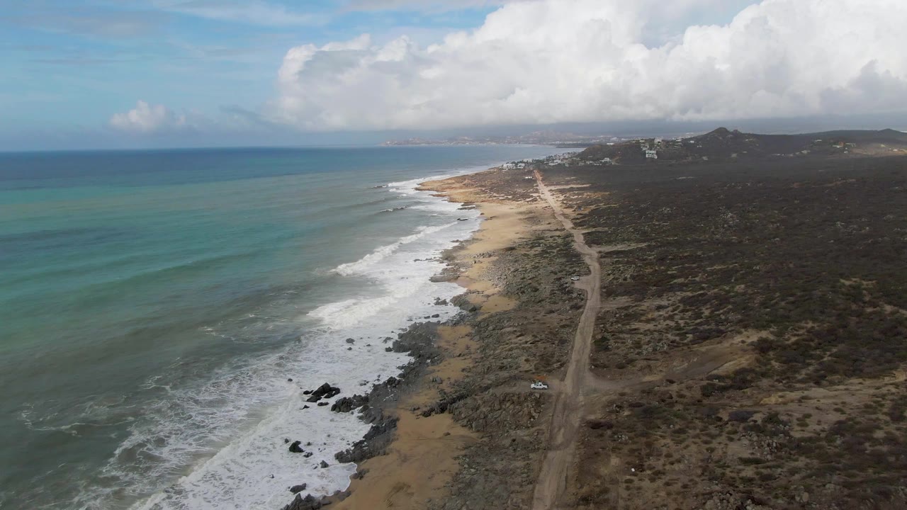 Opening aerial frame over Punta Gorda, Baja California Sur, with teal ocean at left, white surf reaching a rocky sandy shore, a dirt road on the right, and no visible people.