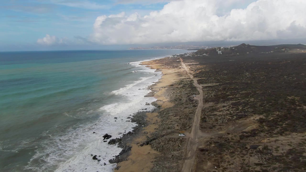 Aerial frame in Punta Gorda, Baja California Sur, showing longer white foam bands along the beach, the coastal road tracking inland from the shore, and no visible people.