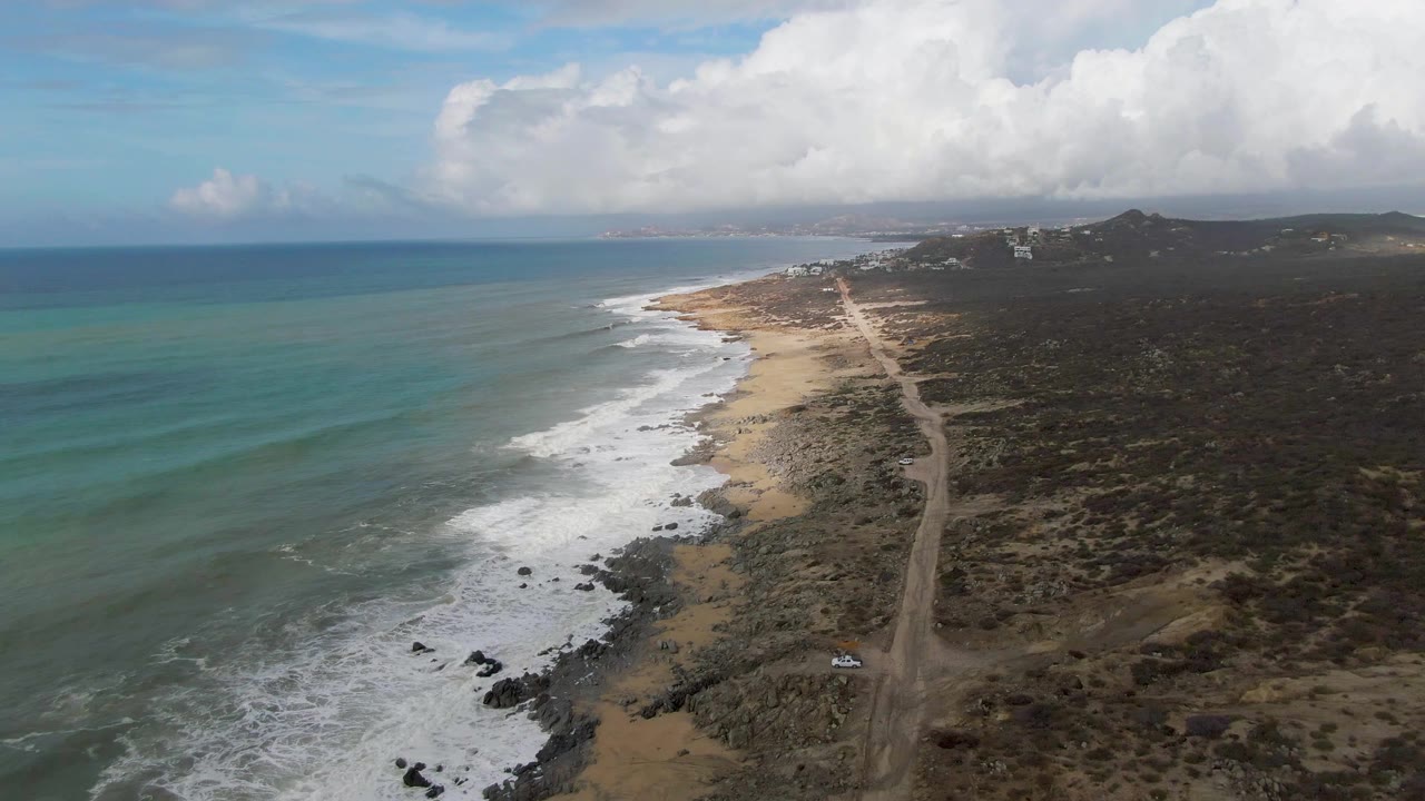 Mid-clip aerial view over Punta Gorda, Baja California Sur, where a small white car is visible near the lower right on the coastal road beside surf and no visible people.