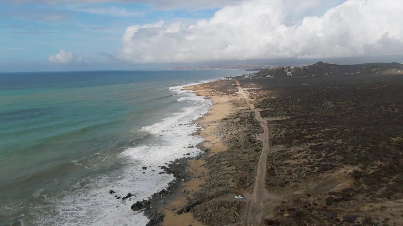 Wide drone view of a beach road, surf, rocks, scrubland, and clouded horizon.